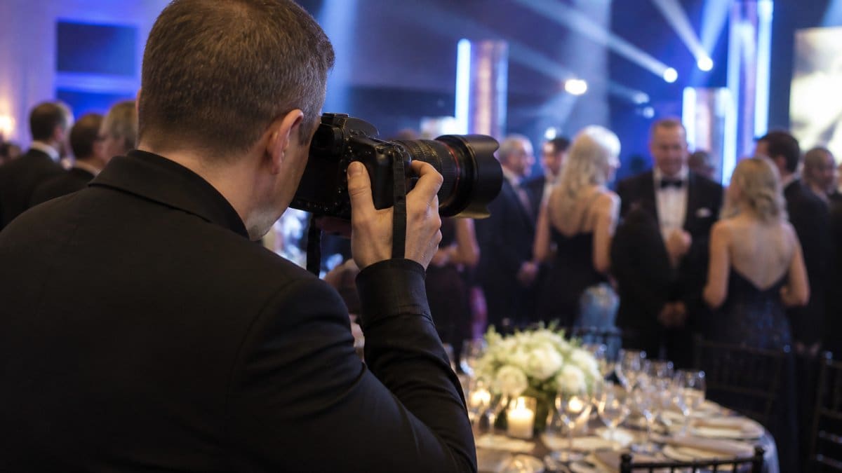 Professional photographer capturing candid moments at a corporate gala, holding a full-frame mirrorless camera with fast lens, elegantly dressed attendees and ambient stage lighting visible in the background